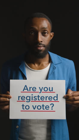 Vertical-Video-Portrait-Of-Vertical-Video-Shot-Of-Man-Holding-Are-You-Registered-To-Vote-Sign-In-Election-Against-Black-Background